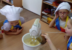 Dziewczynki obserwują dodawanie oleju do miksującego się ciasta.